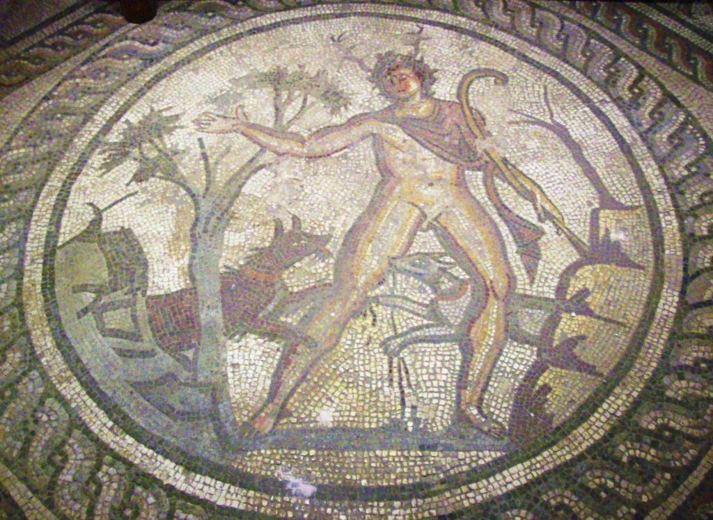 Roman floor mosaic in a circular frame showing Actaeon with small antlers, arms flung out, as hunting dogs close in around him.
