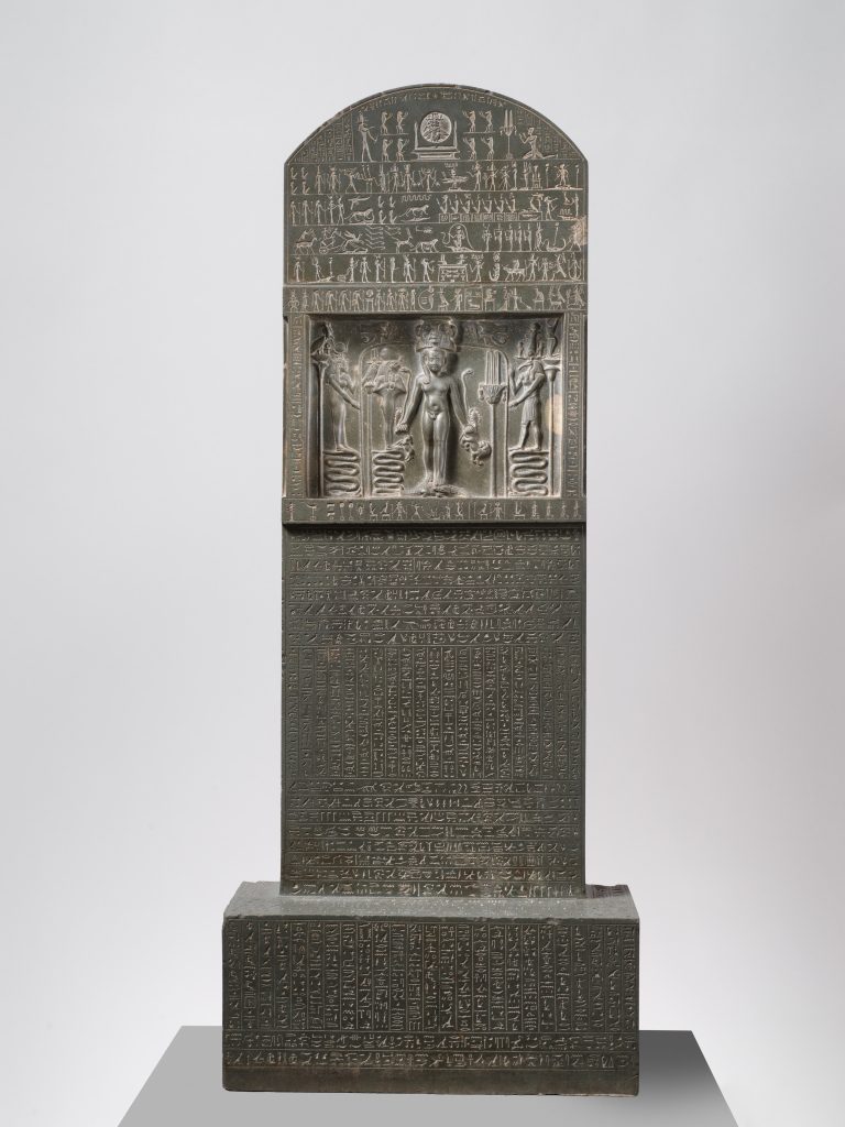Dark stone stela showing Horus standing on crocodiles above rows of hieroglyphic spells.