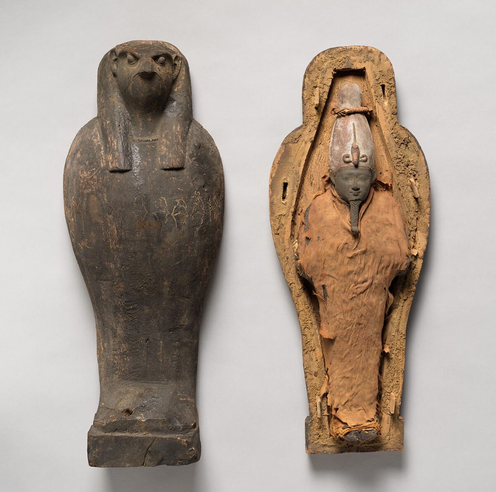 An opened falcon-headed coffin beside its lid, revealing a wrapped symbolic figure with an Osiris mask inside.