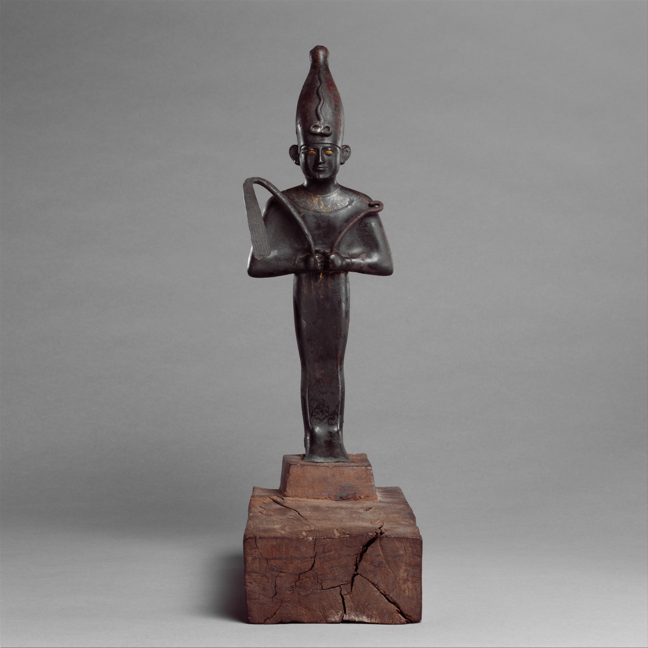 Bronze figure of Osiris wearing a tall crown and holding a crook and flail, mounted on a rectangular wooden base.