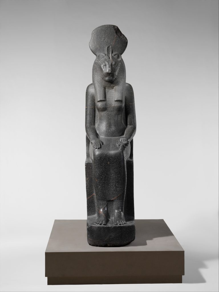Standing lioness-headed Sekhmet statue wearing a sun disk, carved from dark stone and photographed against a pale background.