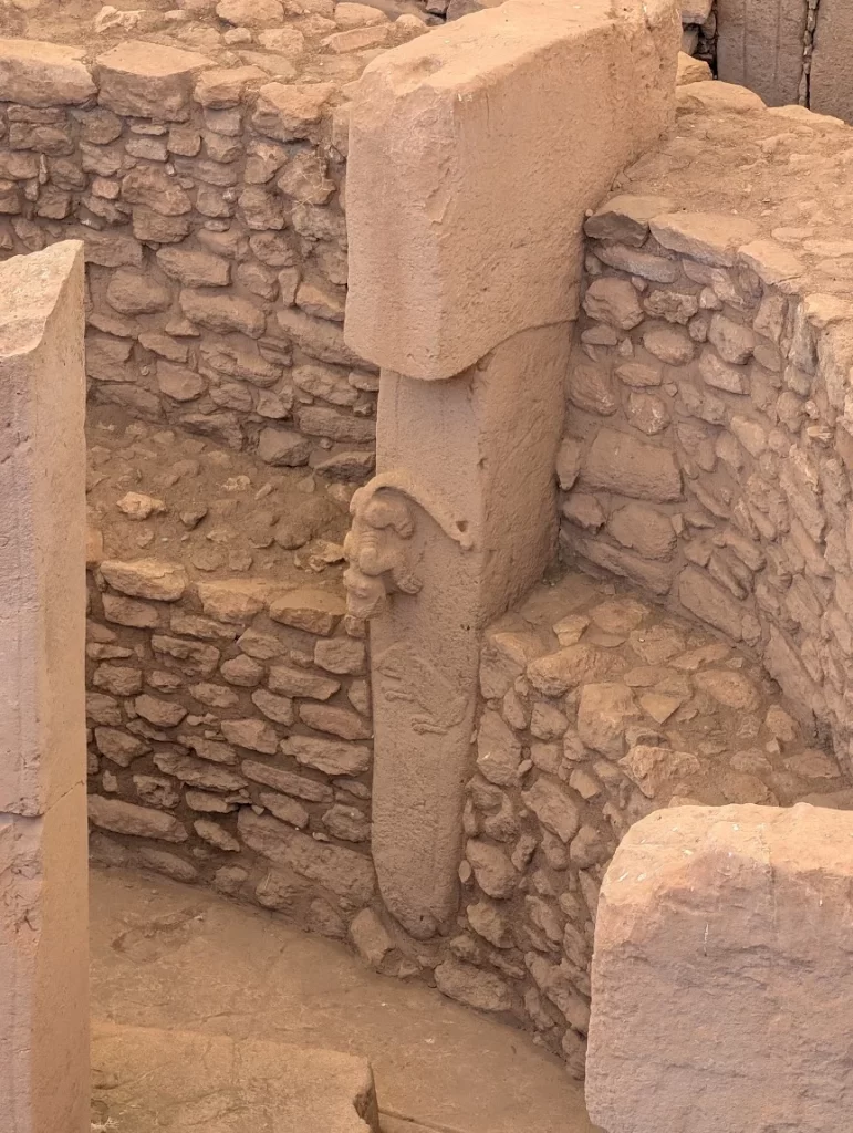 A T-shaped limestone pillar standing against a dry-stone wall. A snarling animal carved in the round climbs head-down on the front of the shaft, and below it the outline of a boar is cut in low relief.