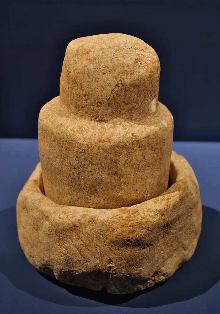 A thick, round-headed limestone pestle standing in a deep, rough stone mortar, photographed against a dark blue background.